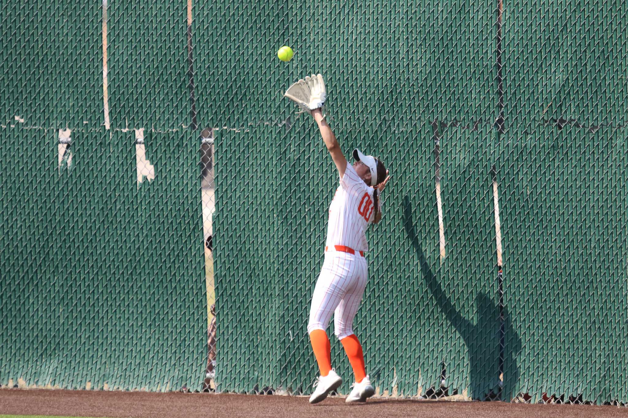 dumas-softball-tyler-anderson-pps-1 | Press Pass Sports Baseball player in white and orange uniform reaches upward with glove to catch a bright green ball near a tall green chain-link fence.