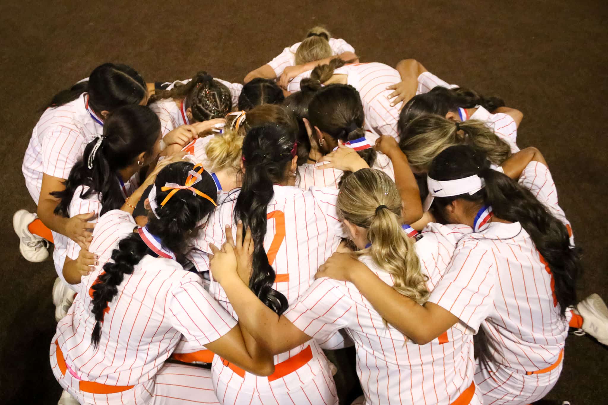 dumas-softball-tyler-anderson-pps-3 | Press Pass Sports Group of athletes in white pinstriped jerseys huddled with arms around each other.