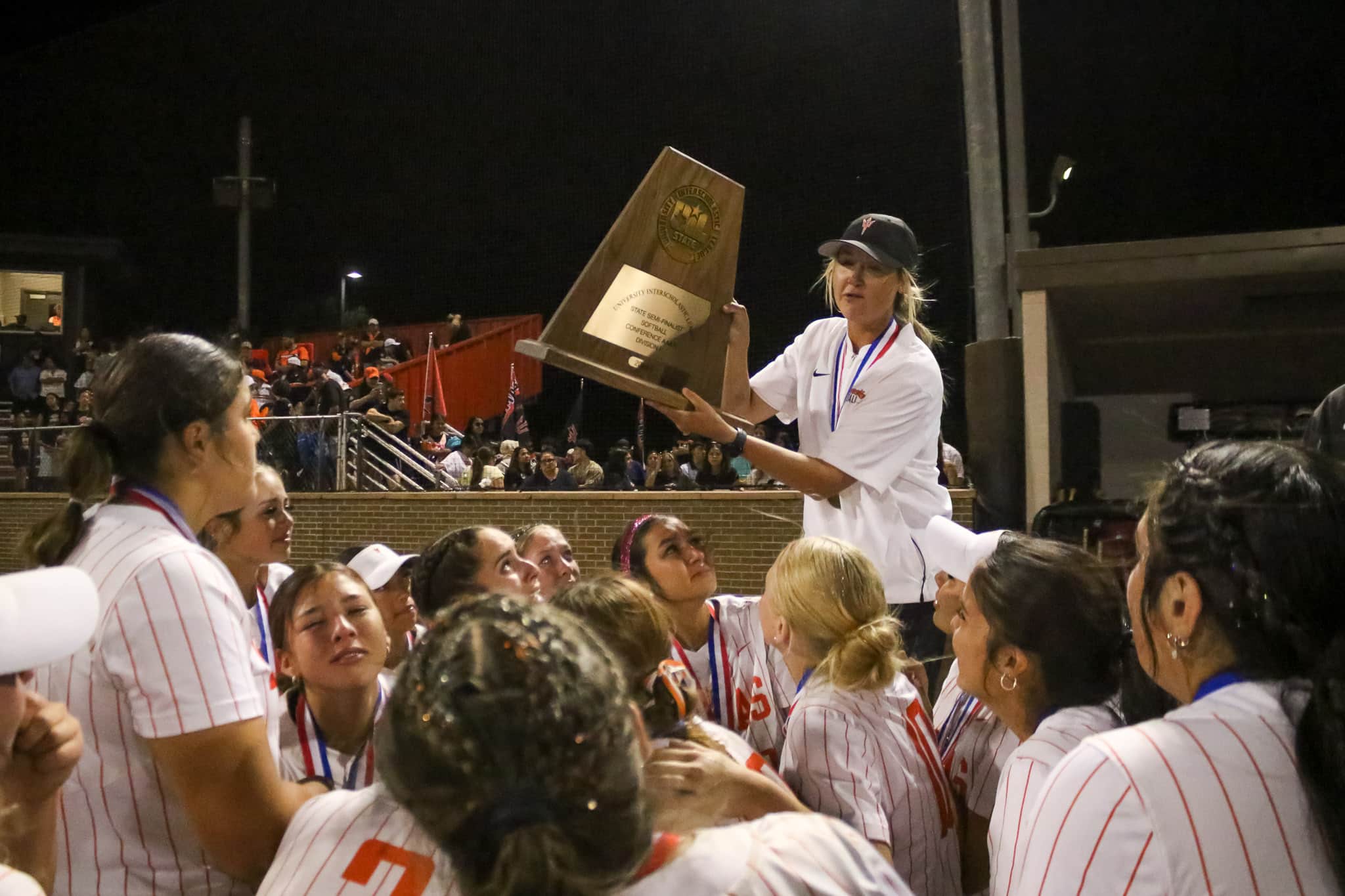 dumas-softball-tyler-anderson-pps-4 | Press Pass Sports Female athlete in a white pinstripe jersey lifts a large wooden trophy as teammates cheer at a nighttime awards celebration on the field