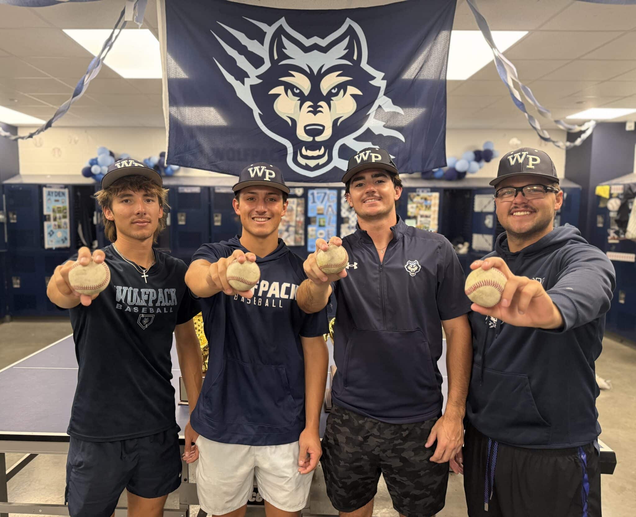IMG_5162 | Press Pass Sports Four male baseball players in navy outfits hold baseballs toward the camera in a gym, with a large wolf logo banner overhead.