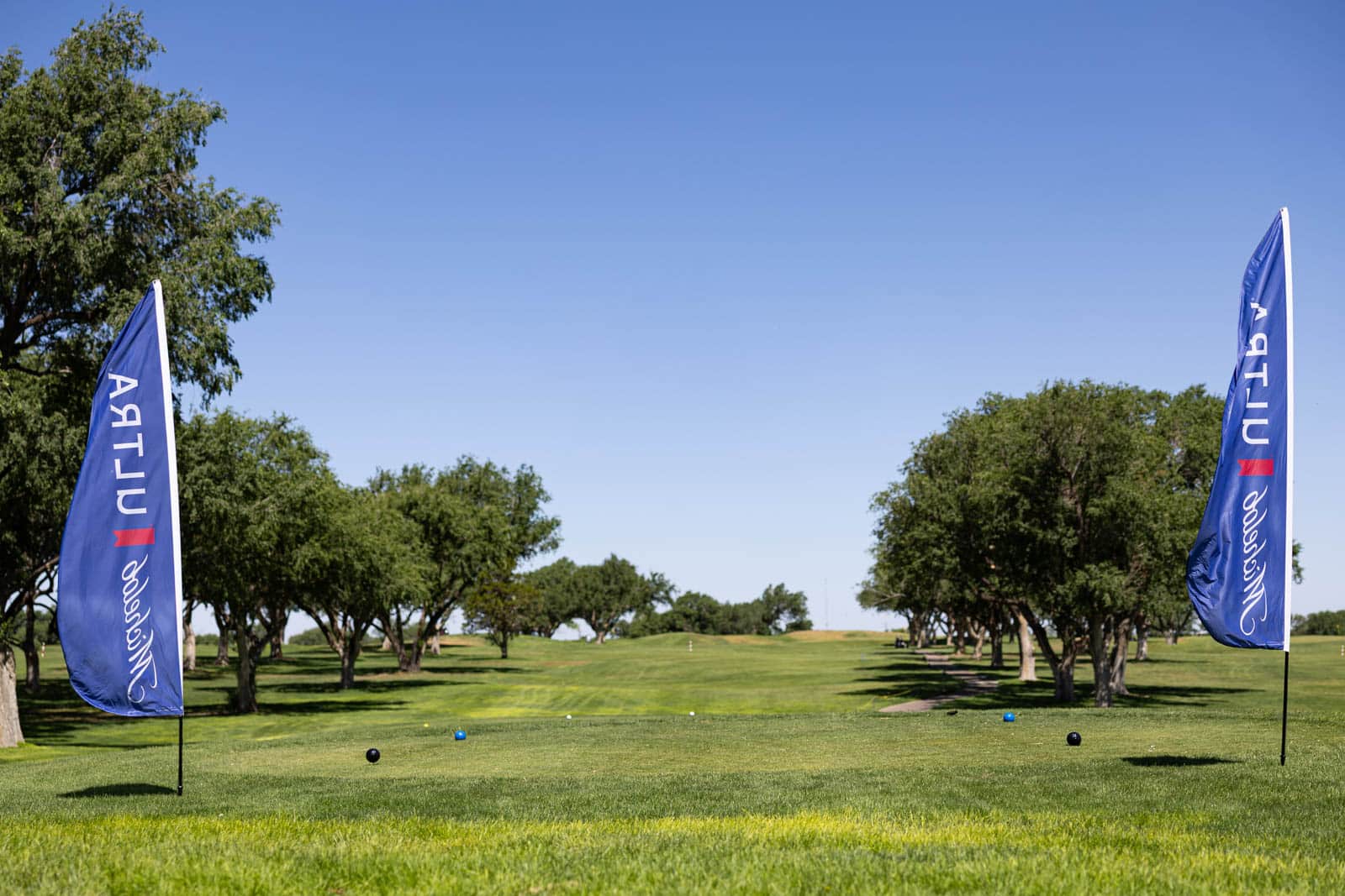 Blue promotional flags stand on either side of a tree-lined golf fairway on a bright, clear day with several golf balls on the tee.