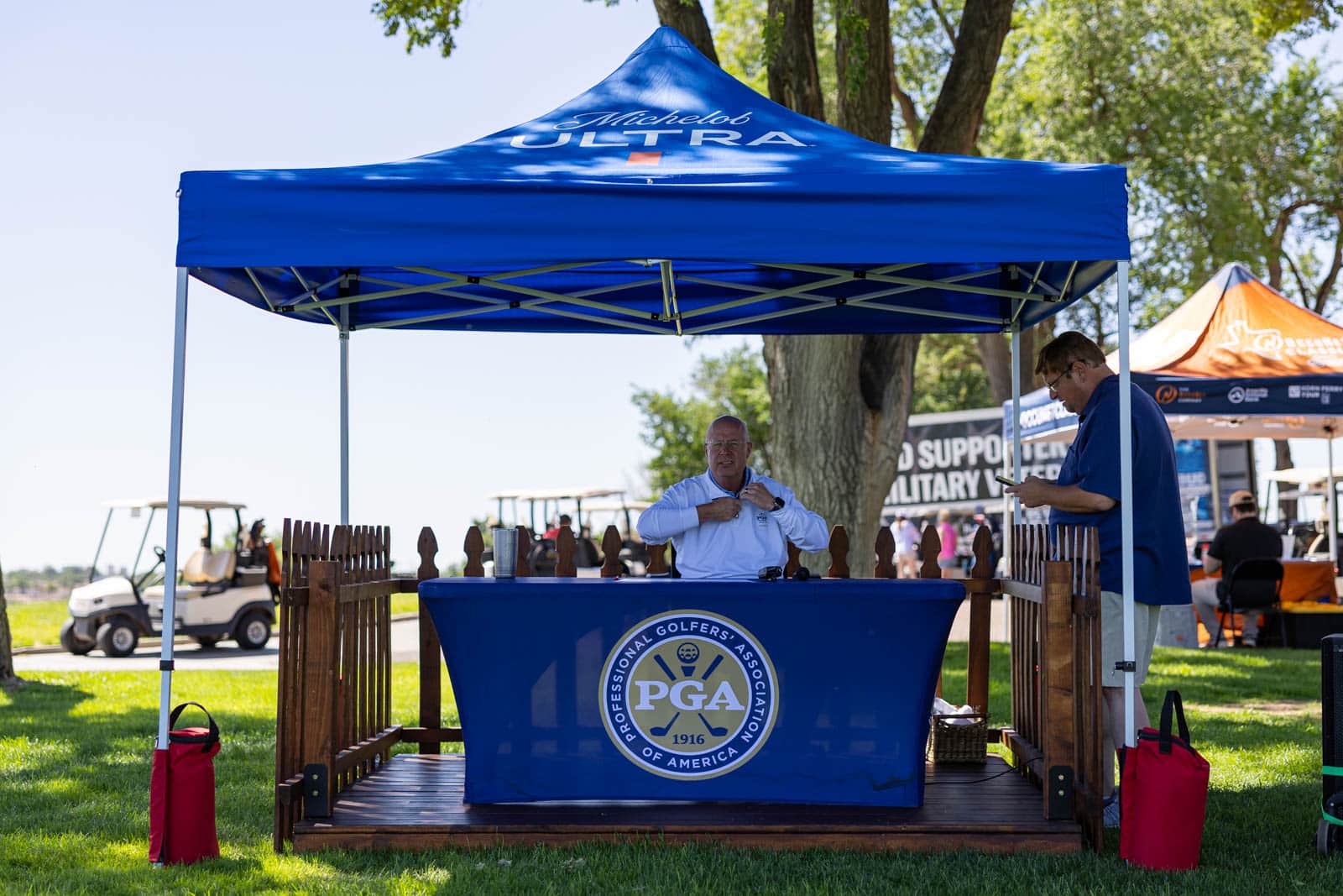 Two men at a PGA booth under a blue canopy at an outdoor golf event with attendees in the background and a golf cart nearby.