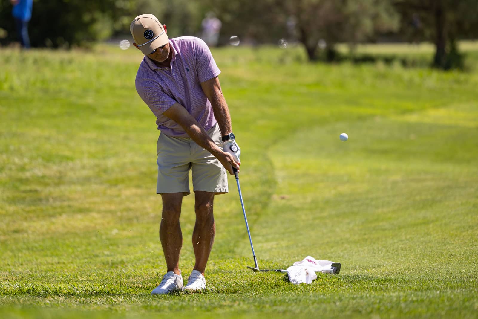 Man wearing a pink polo and beige shorts swings a golf club on a sunny golf course; ball is in midair nearby