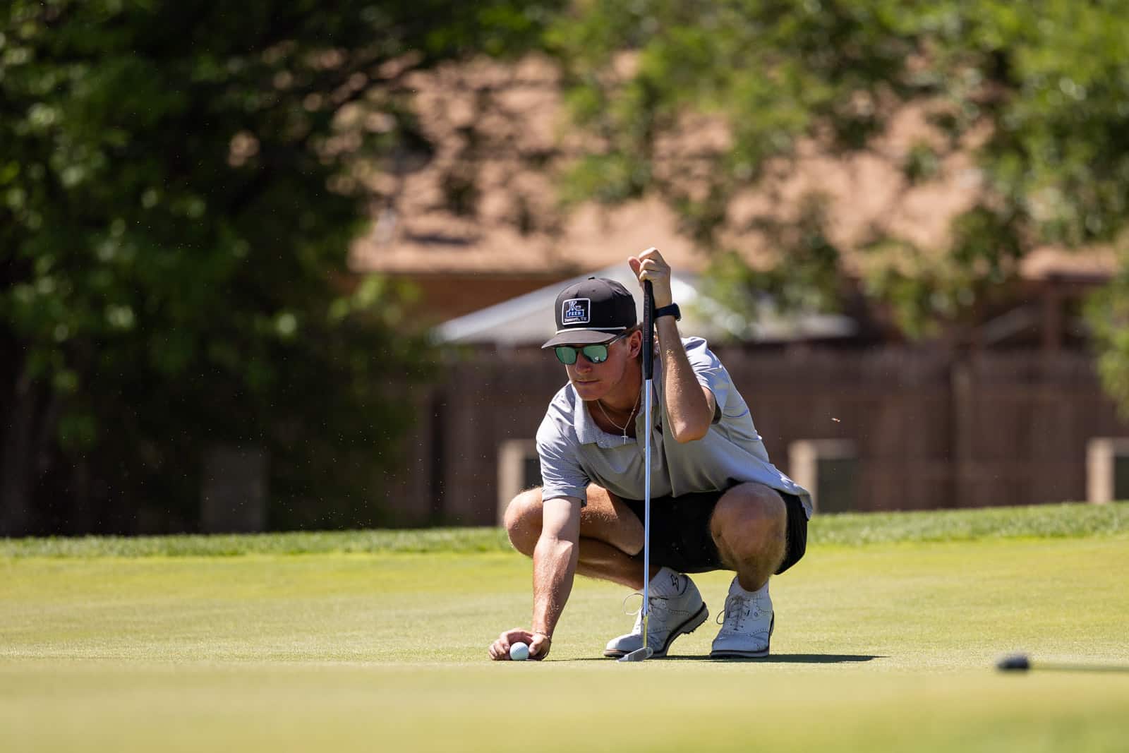 Male golfer crouches on the green, lining up a putt with a putter while wearing a cap and sunglasses