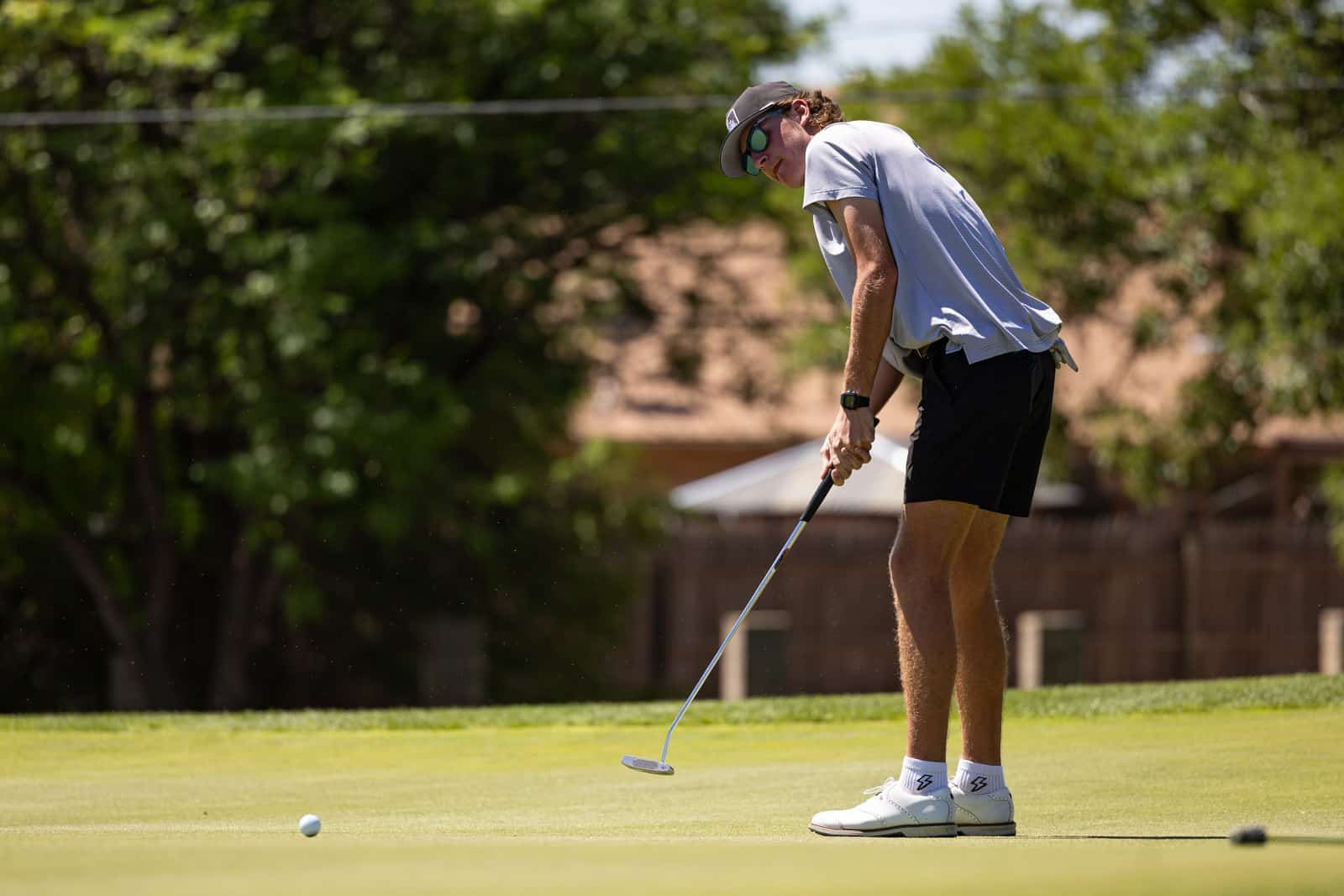 A man wearing a cap and sunglasses lines up a putt on a putting green, preparing to hit the ball.
