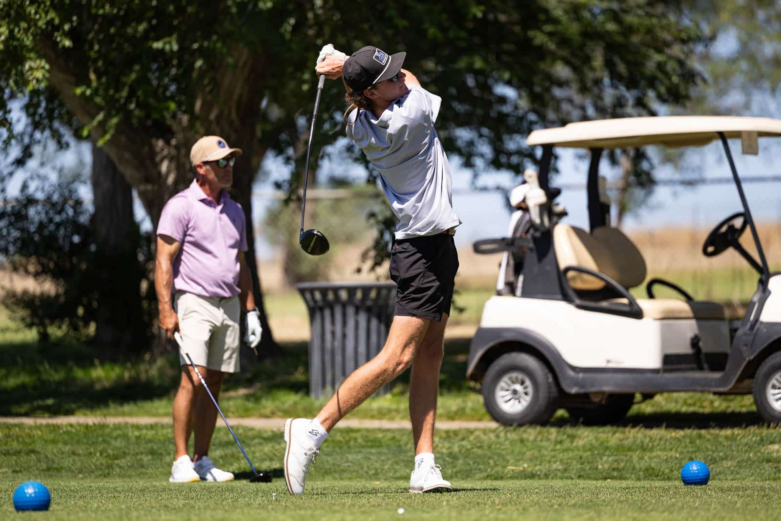 Golfer in a gray shirt and black shorts mid-swing on a sunny course, with a spectator watching and a golf cart in the background.