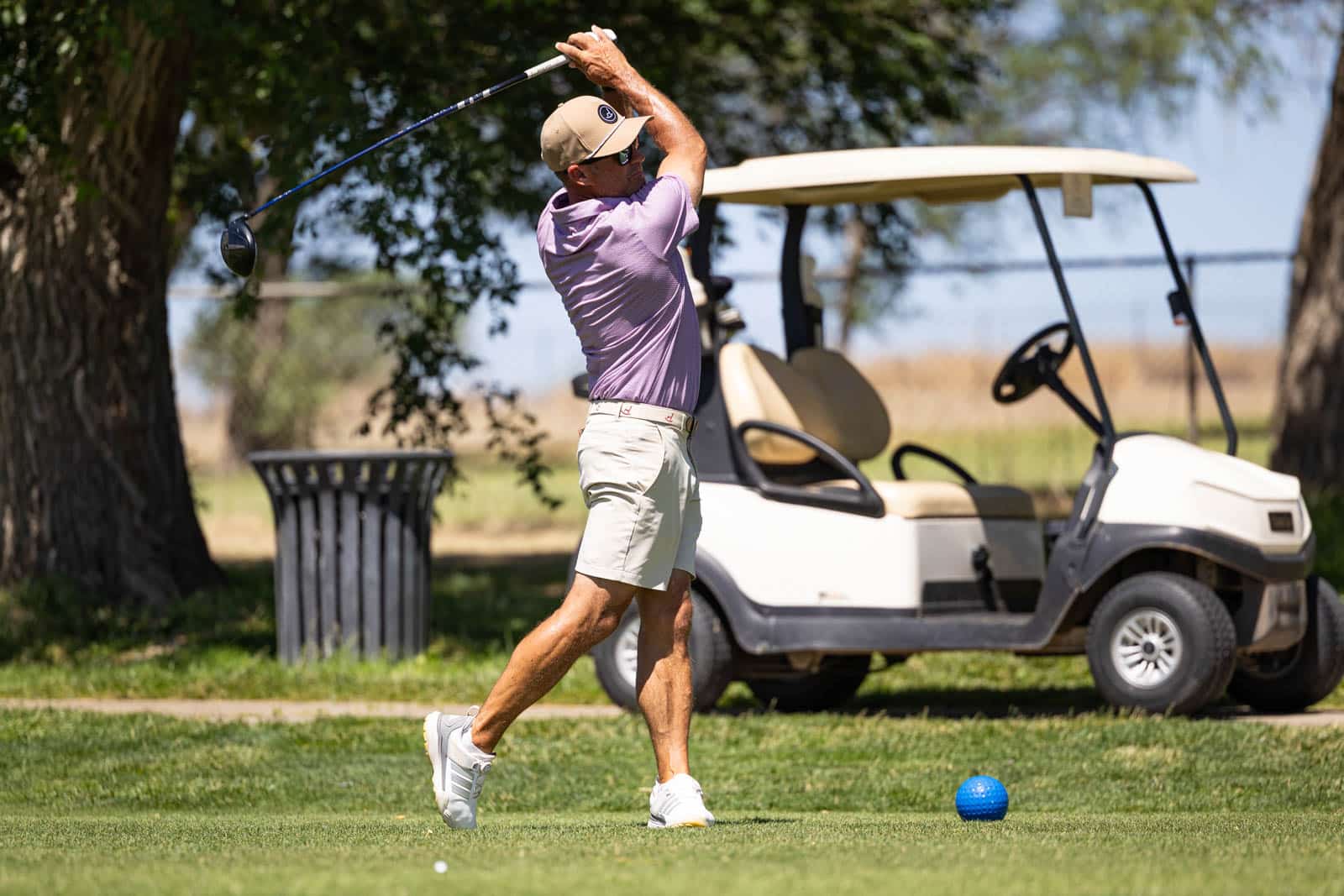 Man in a purple polo and beige shorts mid-swing on a sunny golf course; a beige golf cart is behind him.