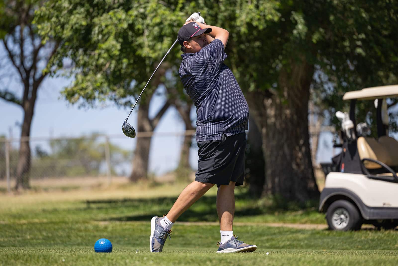 Golfer in mid-swing on a sunny course, wearing a navy polka-dot shirt and black shorts, with a blue ball on the grass nearby.