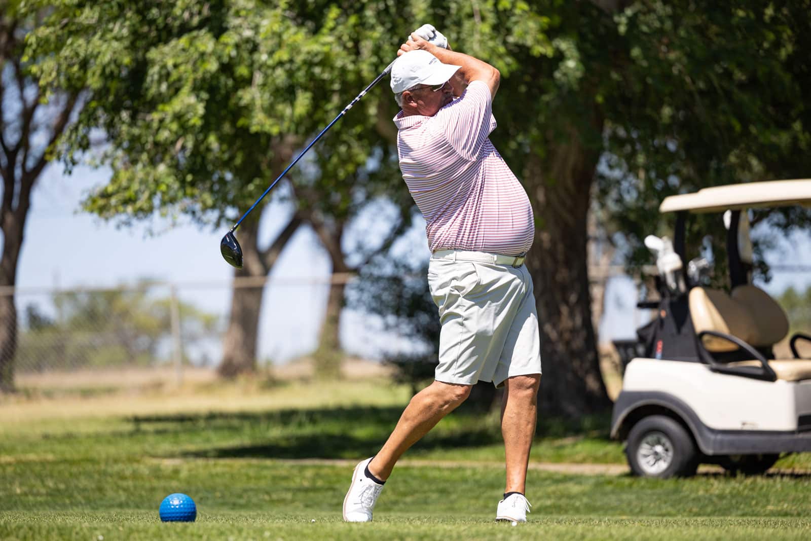 Older man in a striped polo shirt and white cap mid-swing on a golf course, with a blue golf ball nearby and a golf cart in the background