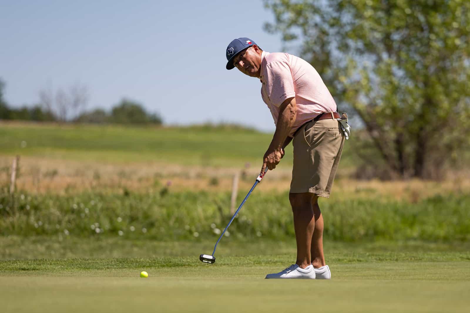 Golfer bending to putt on the green, lining up a shot with a yellow ball nearby on a sunny course.