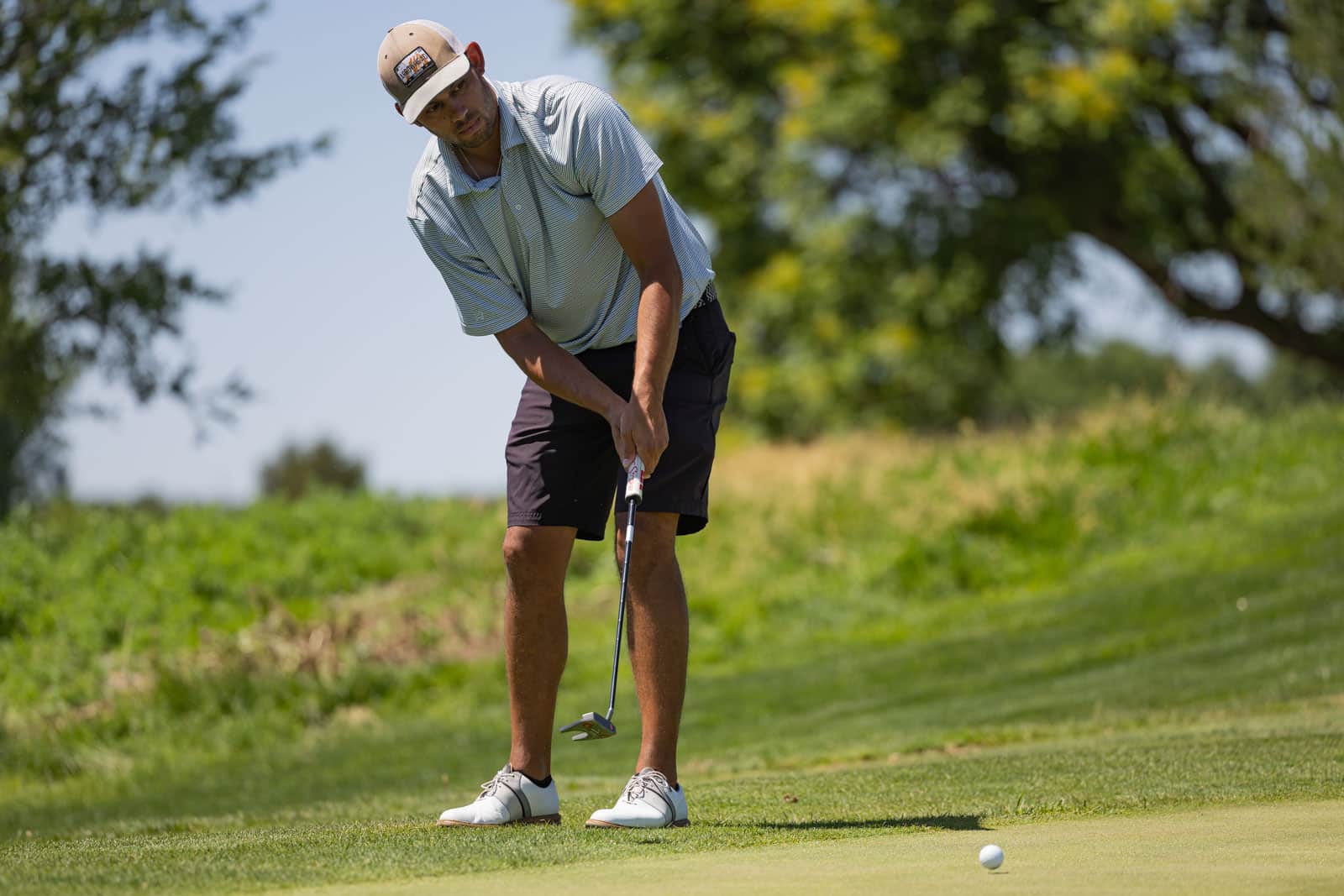 Golfer lining up a putt on a sunlit green, wearing a striped shirt and beige cap, with trees in the background.