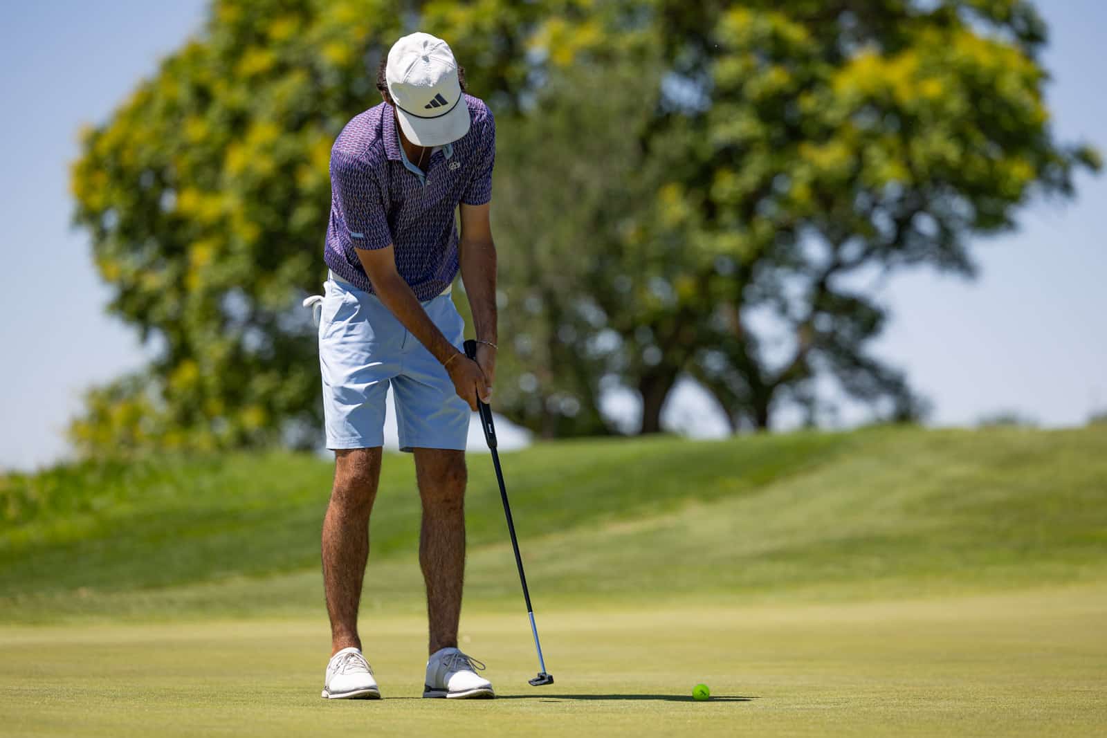 Male golfer putting on a sunny golf course green, preparing to strike the ball with a putter nearby.