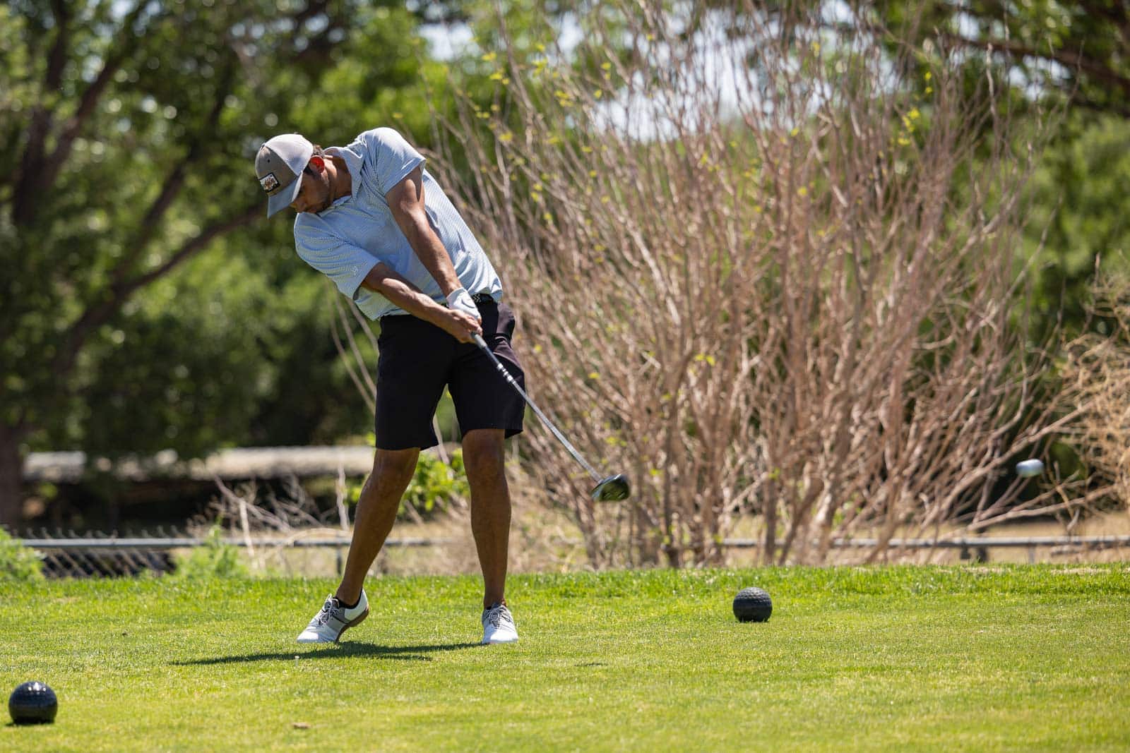 Golfer in a light blue shirt and cap mid-swing on a sunny golf course, ball teed up ahead of him.