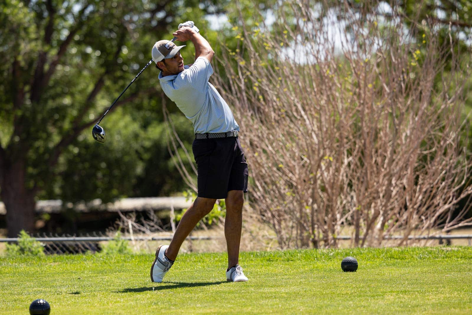 Golfer mid-swing on a sunny course, wearing a cap, light blue polo, and black shorts.