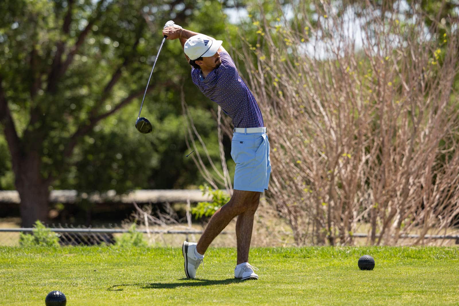 Golfer in a blue patterned shirt and light blue shorts, mid-swing with a driver on a sunny golf course.
