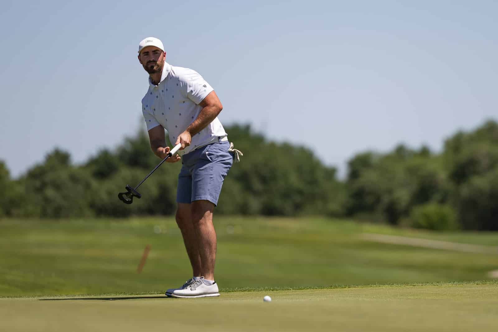 Male golfer wearing white polo and cap lining up a putt on a green, ball nearby with a fairway and trees in the background