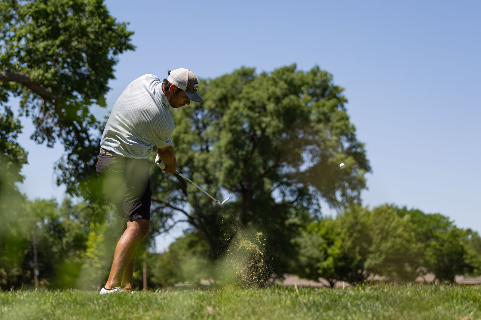 Golfer in a cap and light shirt mid-swing on a sunny golf course, ball in flight.