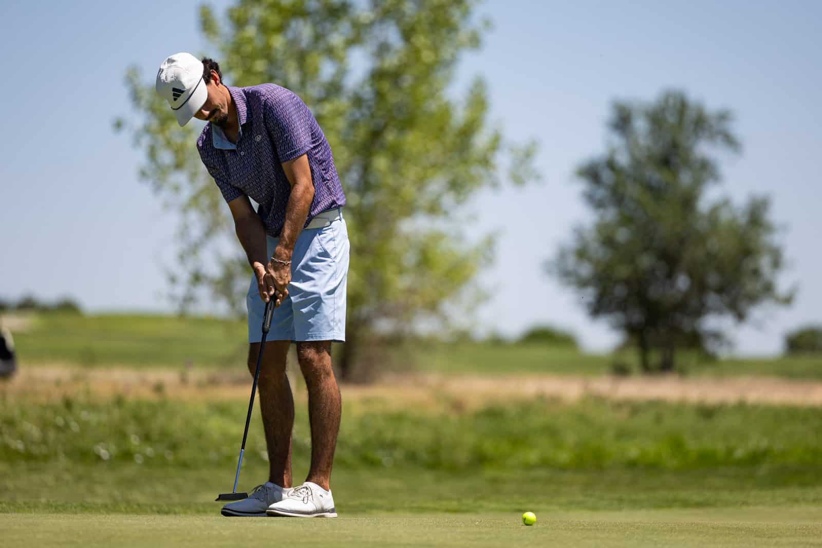 Golfer in a white cap putting on a green fairway with a golf ball nearby, focused stance on a sunny day.