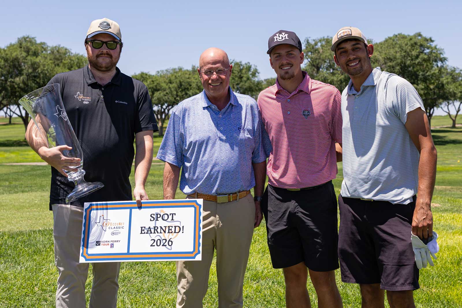 Four men on a sunny golf course pose together; one holds a tall glass trophy, another holds a sign reading “Spot Earned! 2026.”