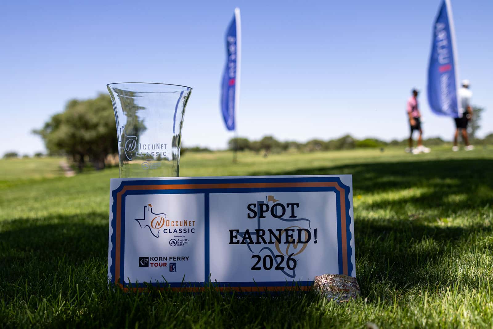 Clear glass trophy beside a sign reading 'Spot Earned! 2026' on a sunny golf course.