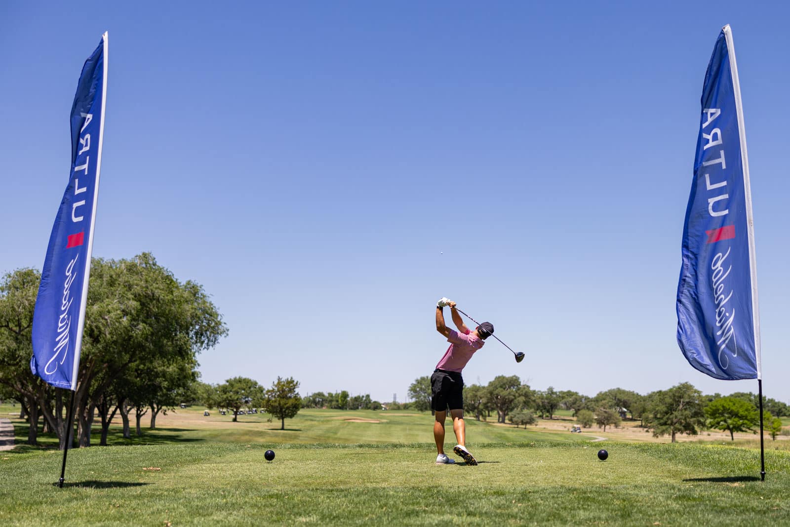 Golfer in a pink shirt mid-swing on a sunny golf course, with tall blue sponsor banners on both sides.