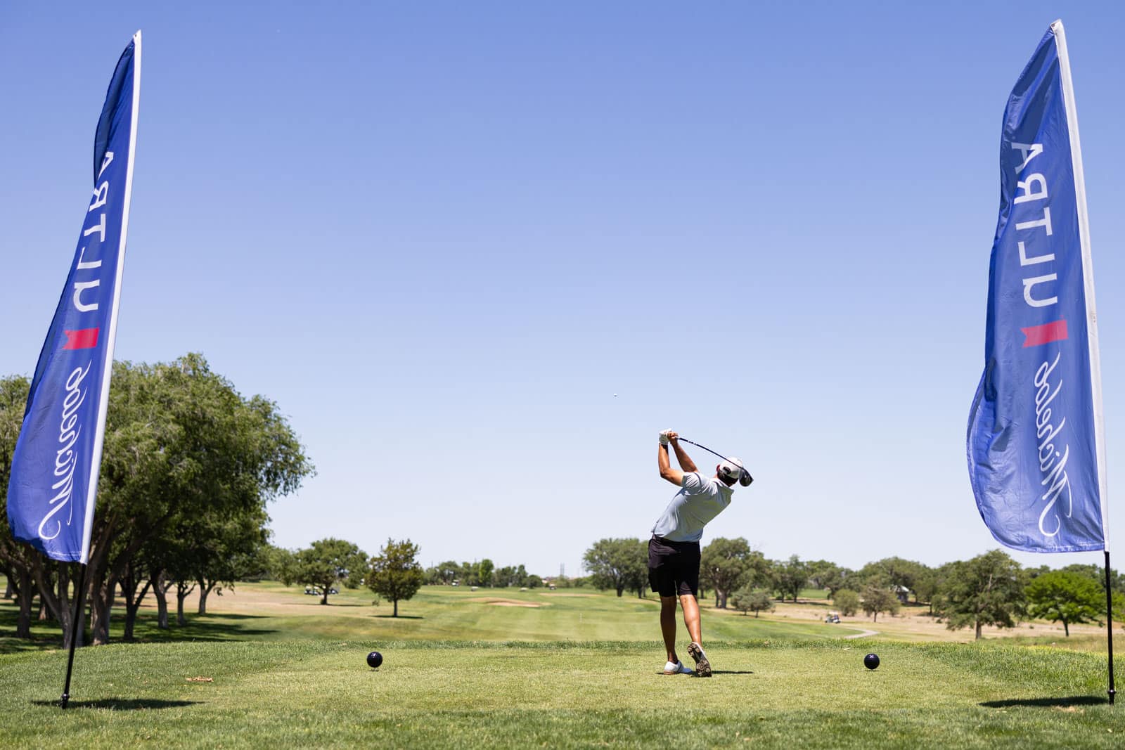 A golfer mid-swing on a tee between tall blue sponsor flags with a clear blue sky and green course beyond.