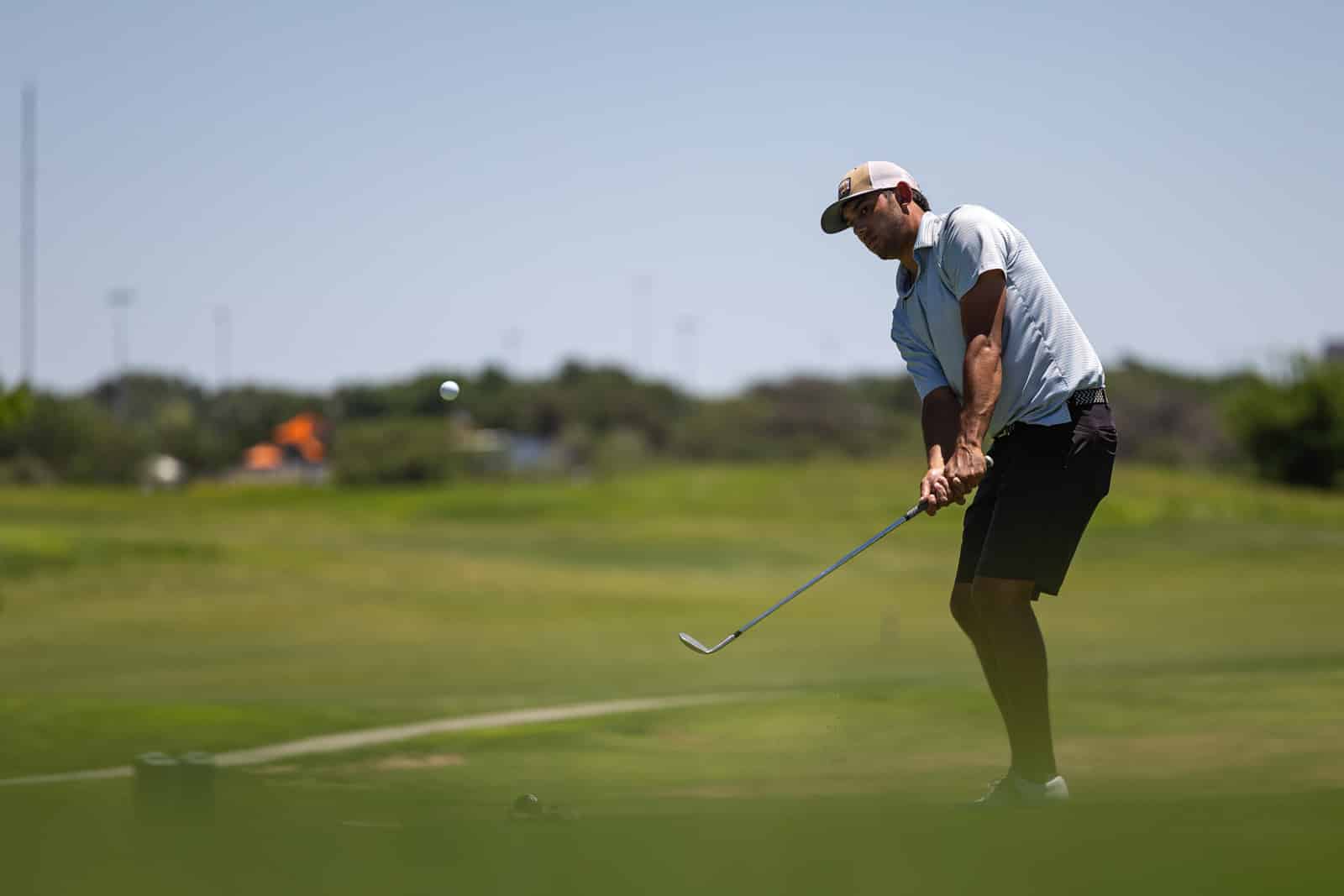Golfer in a striped polo and cap swings a club on a bright, outdoor golf course as the ball rises mid‑flight.