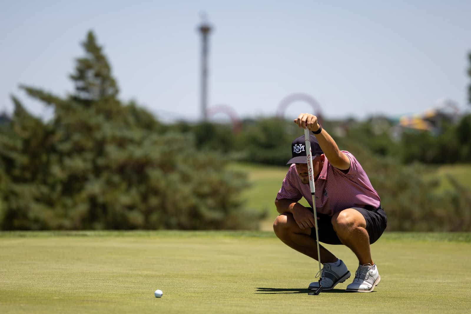 Golfer crouching on the green, lining up a short putt with a putter toward a nearby ball on the grass on a sunny day.