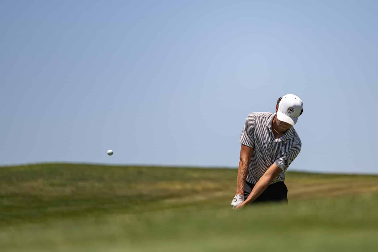 Male golfer in a striped polo and white cap swinging on a sunny golf course with a ball in mid-air against a blue sky.