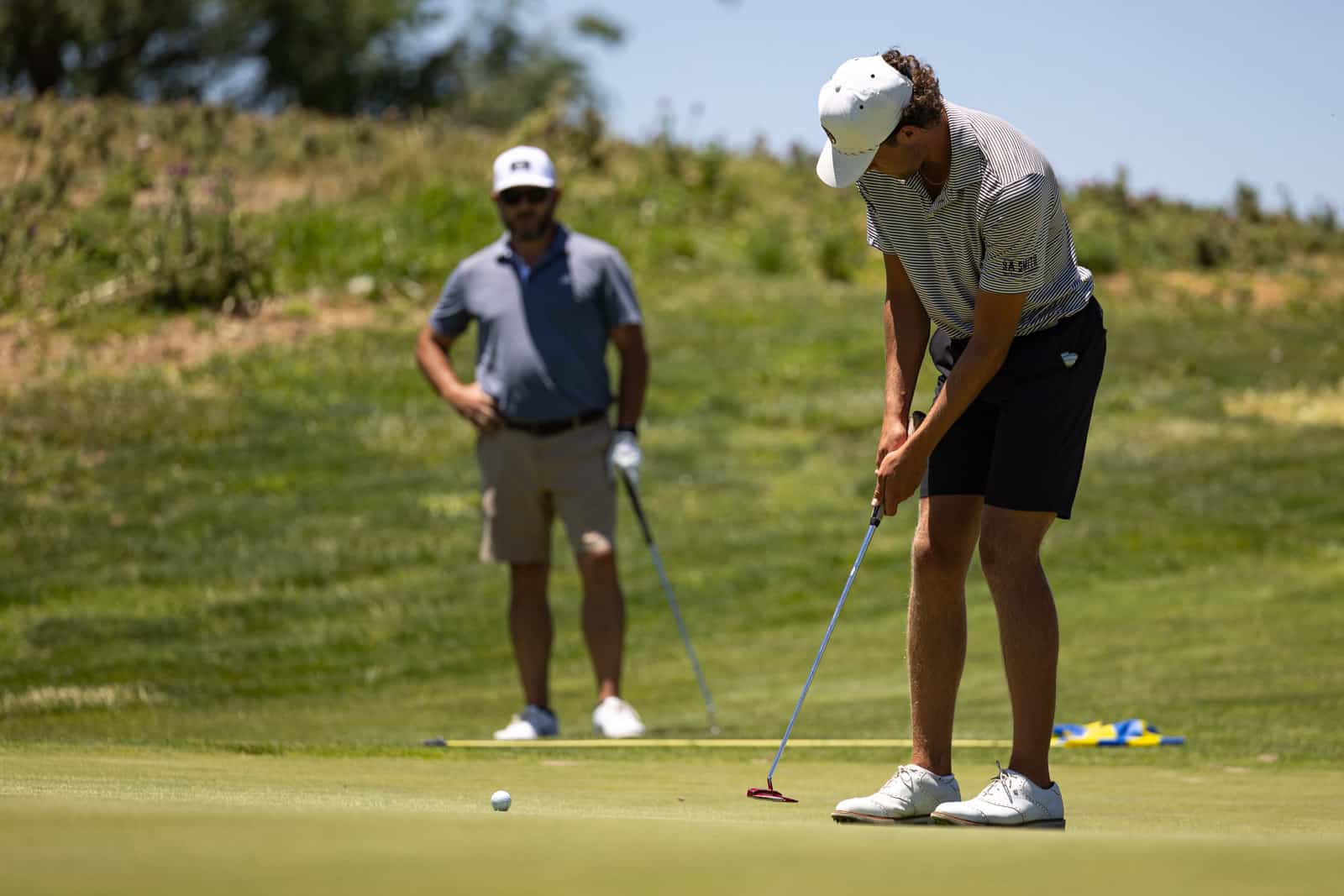 Two men on a sunny golf course: one putts near the hole while the other watches in the background.