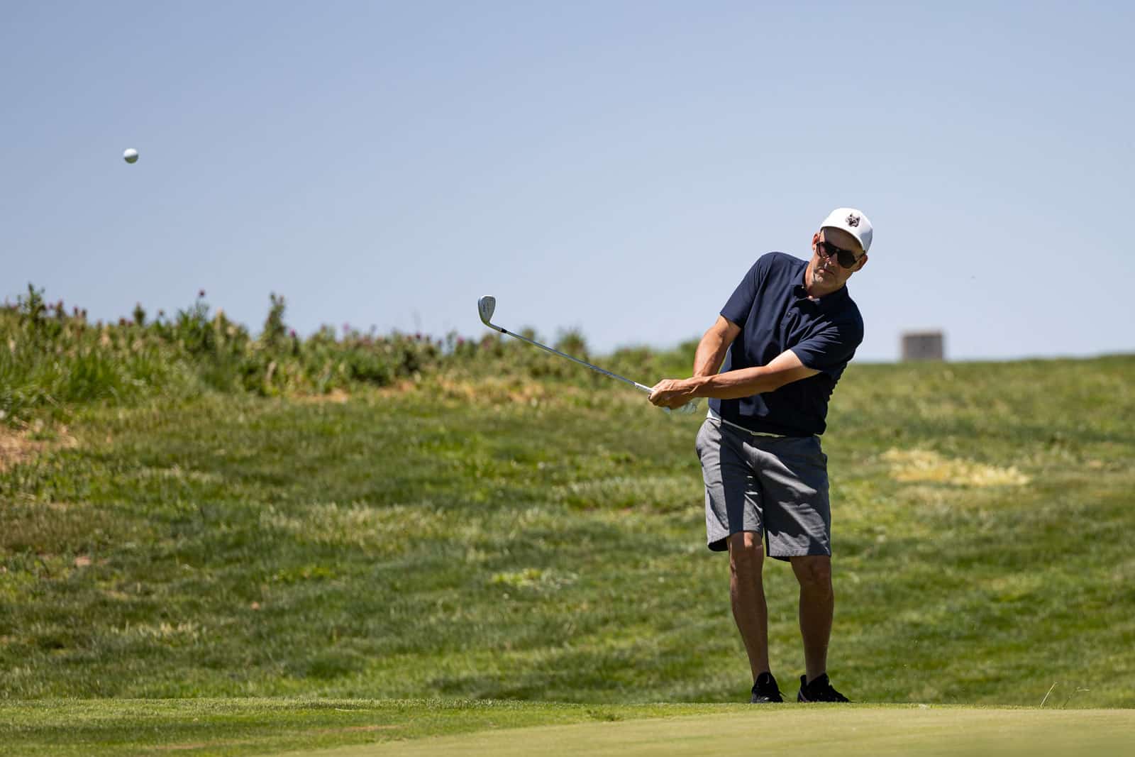 Man in a navy polo and gray shorts mid-swing on a sunny golf course, hitting a shot toward the ball in flight.
