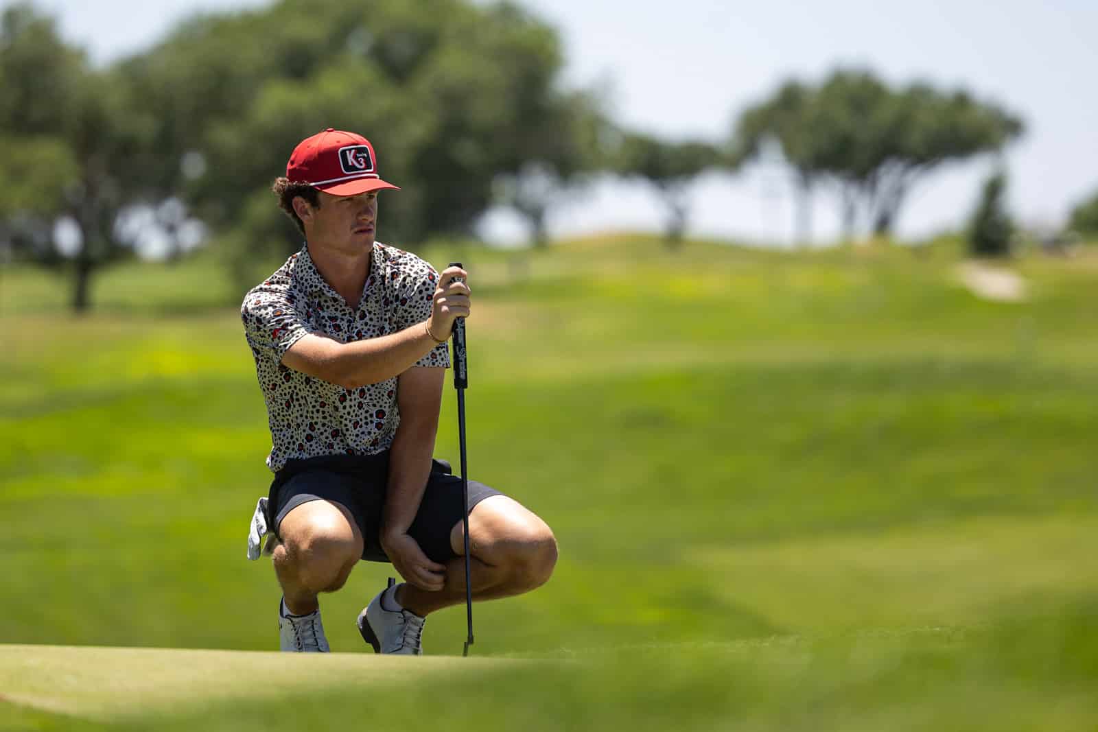 Male golfer kneeling on the putting green, holding a putter and wearing a red cap and patterned shirt.