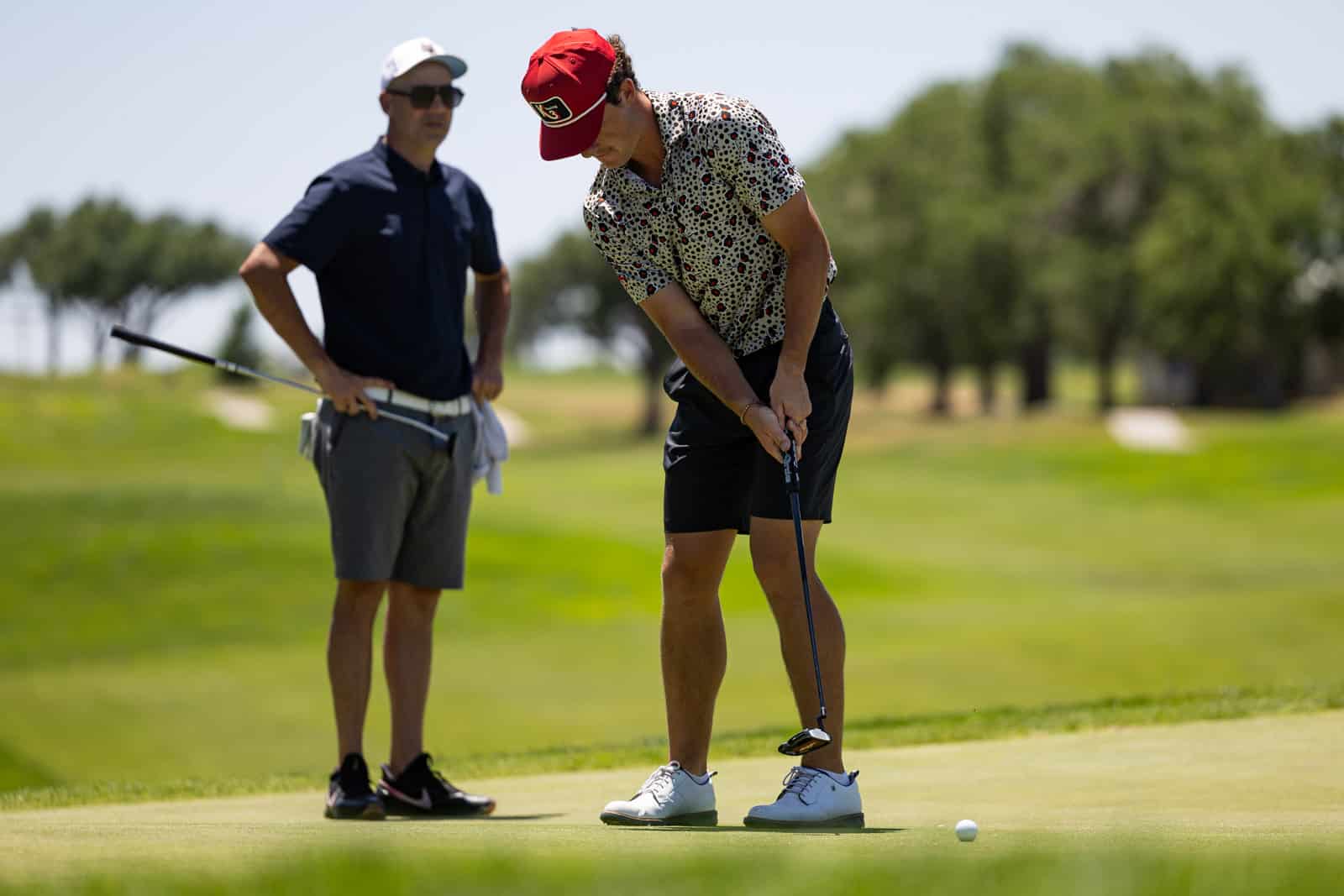 Golfer lining up a putt on a sunny green while a second player watches in the background.