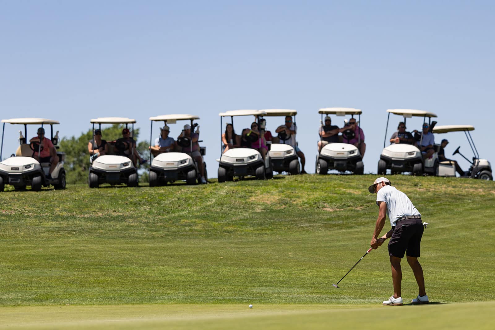 Golfer lining up a putt on the green with a row of golf carts and spectators behind him on a sunny day