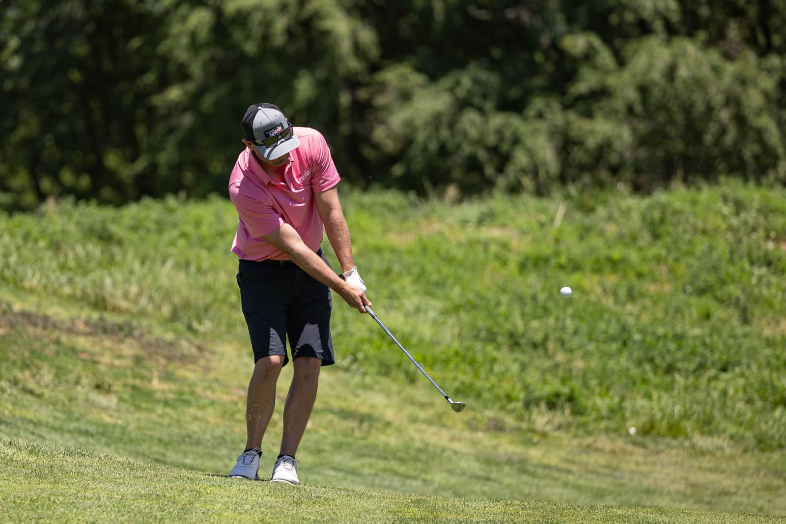 Golfer in a pink polo shirt and cap swinging a club on a sunny, grassy golf course.