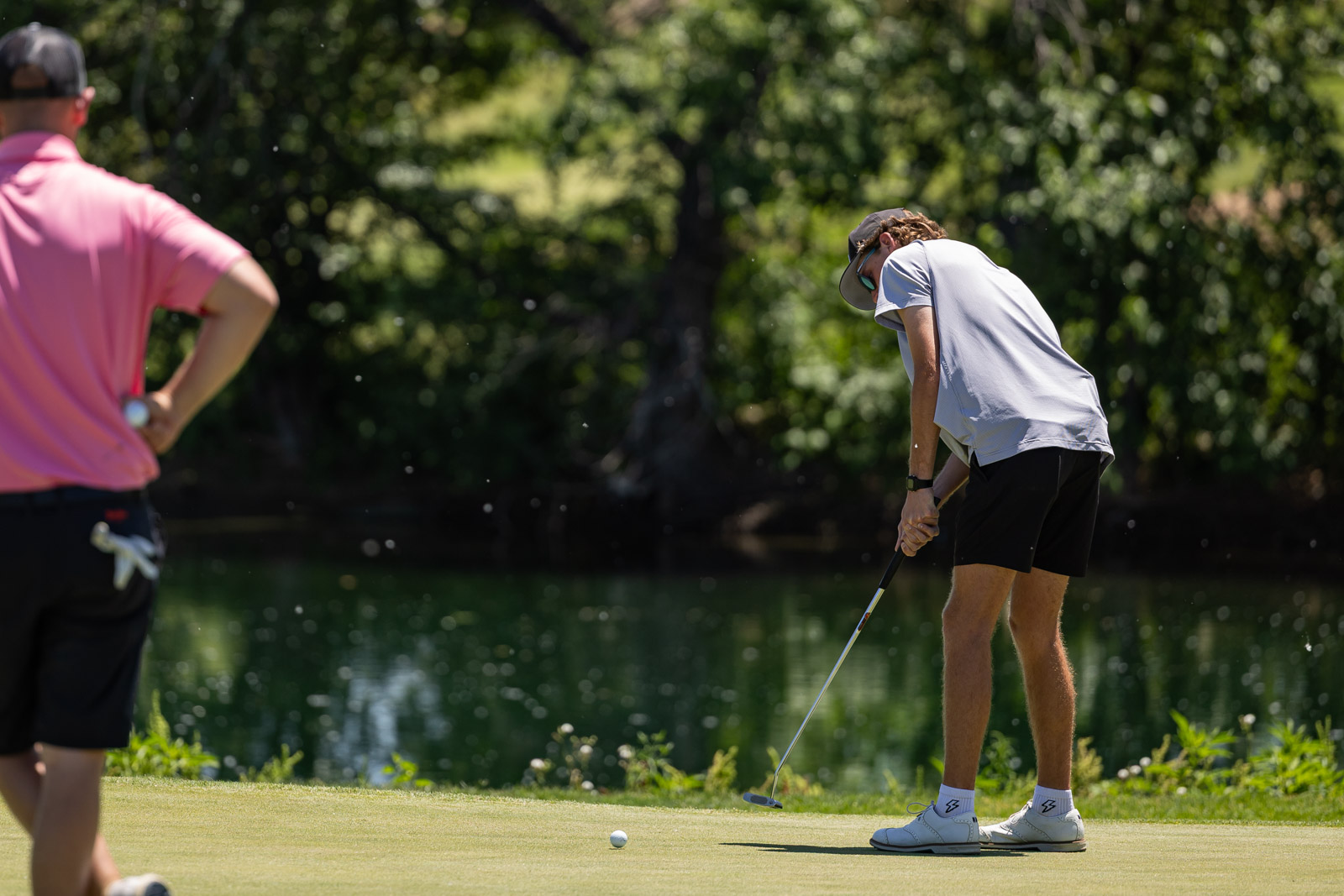 Golfer in a gray shirt and black shorts putting on a green by a pond, while another player in a pink shirt watches to the left.