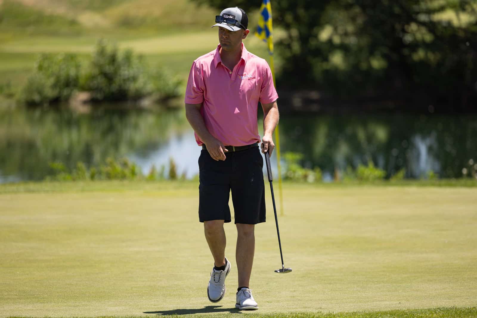 Golfer in a pink polo and black shorts walking on the putting green, holding a putter near a yellow flag with a pond and trees in the background