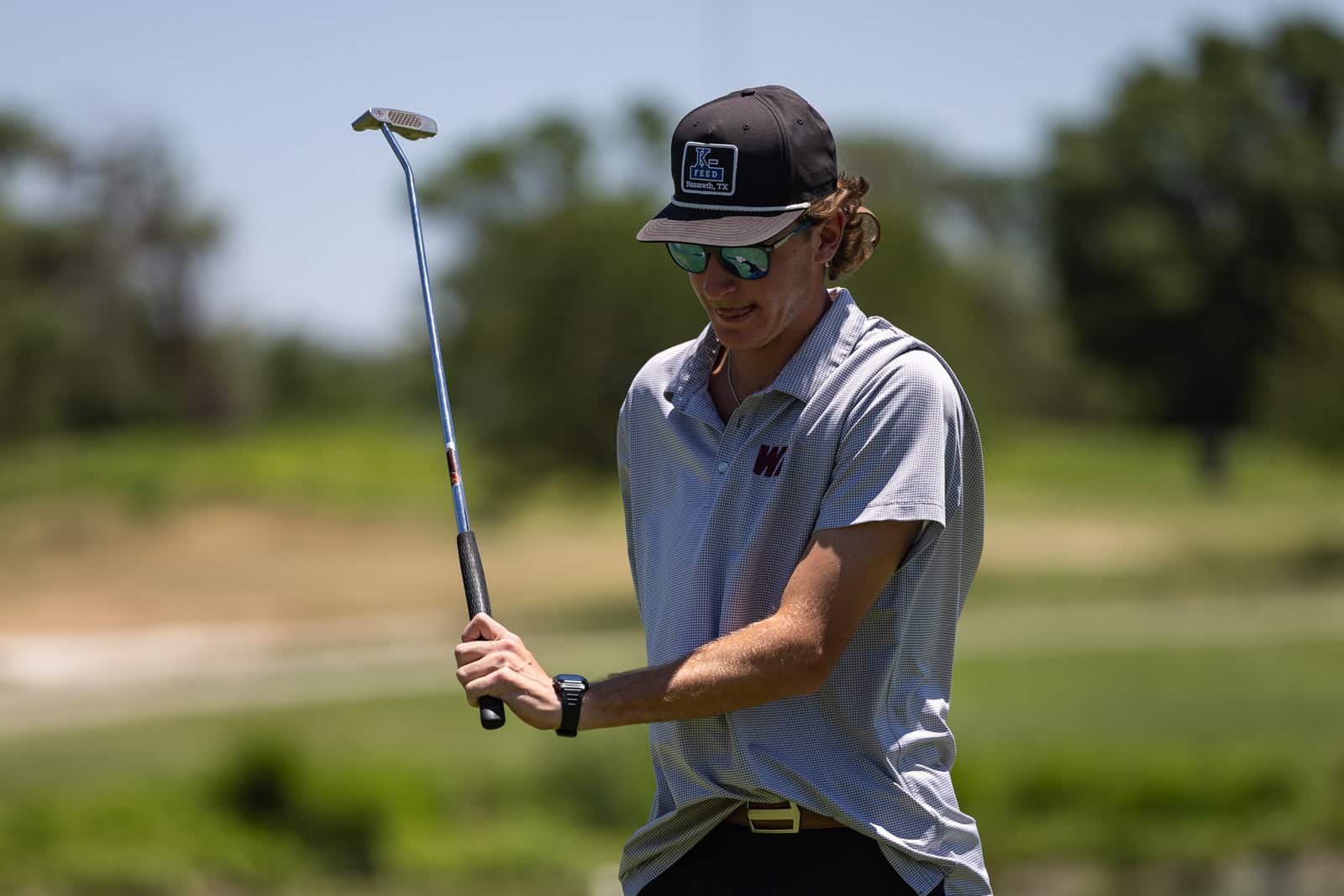 Golfer in a gray polo, black cap and sunglasses holding a club mid-swing on a sunny golf course.