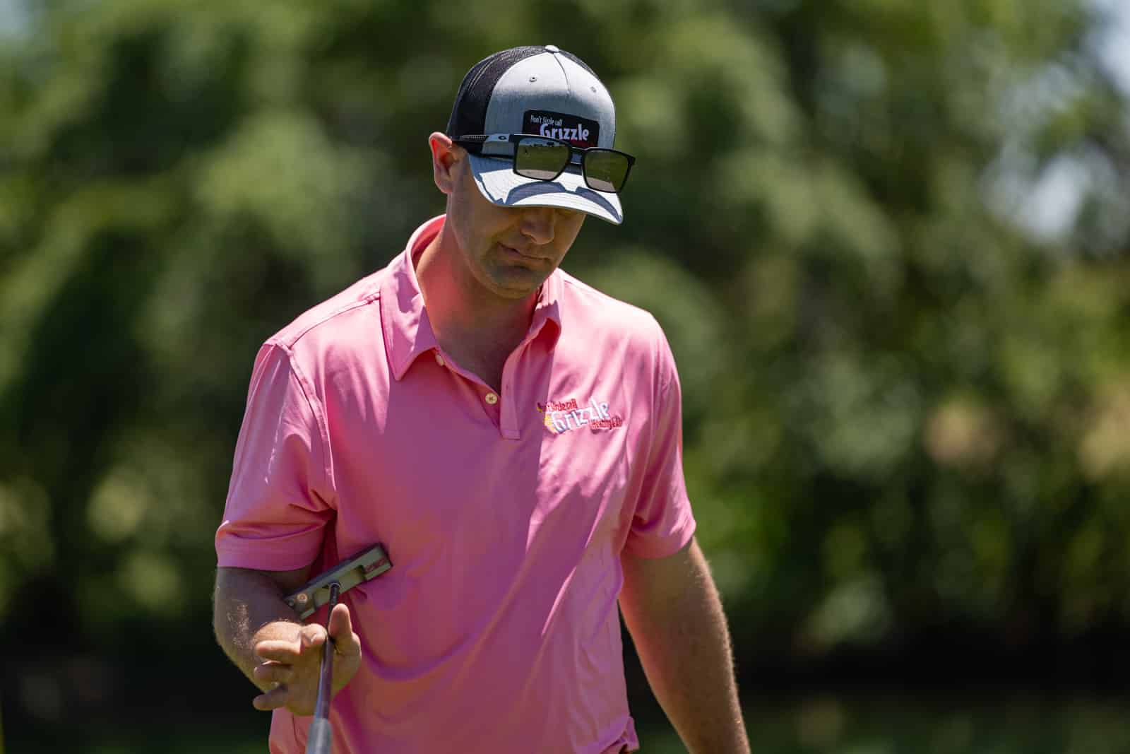 Man in a pink polo shirt and trucker cap with sunglasses on top, holding a golf club outdoors.
