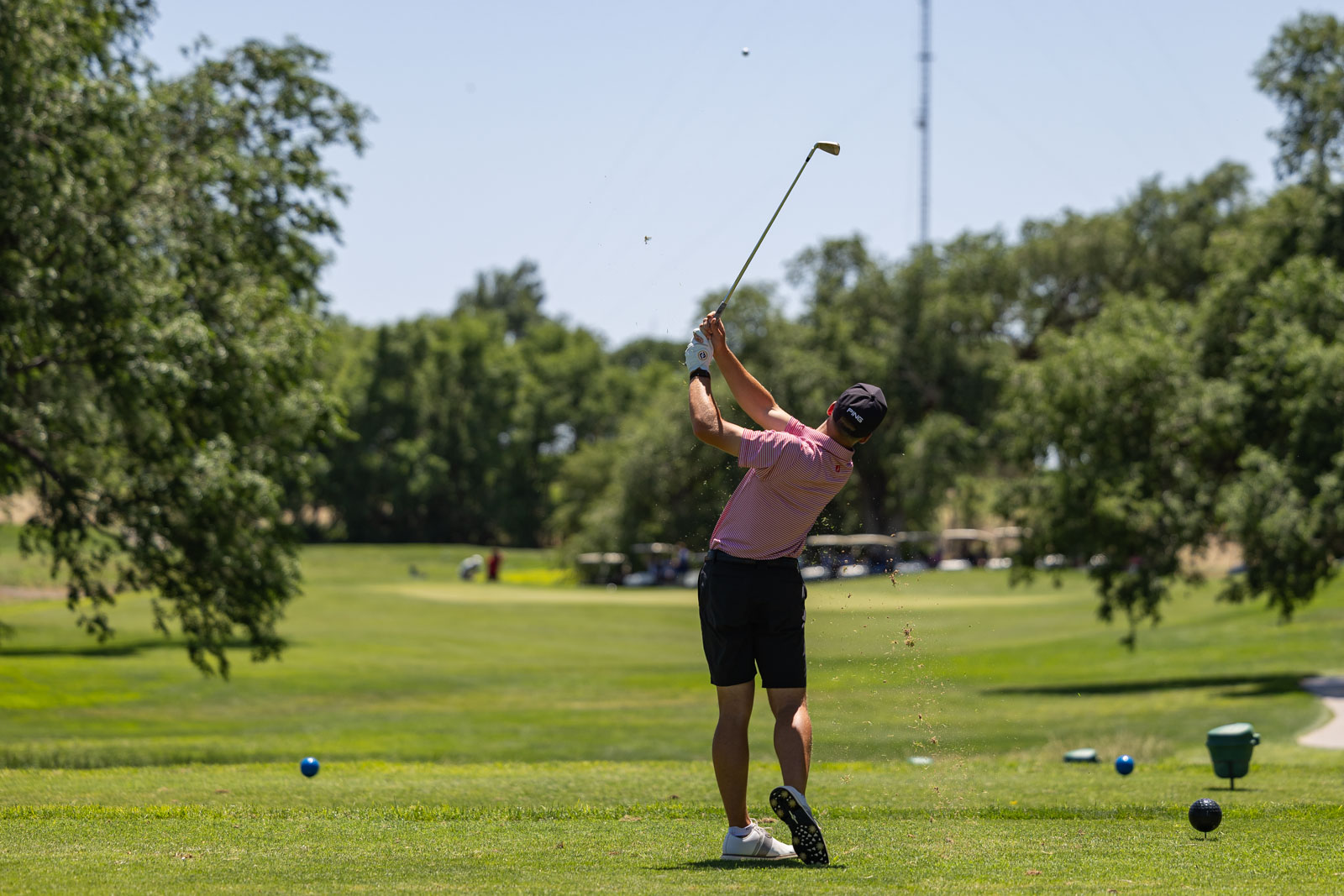 Golfer in a red striped shirt mid-swing on a sunny golf course with trees and green grass in the background.