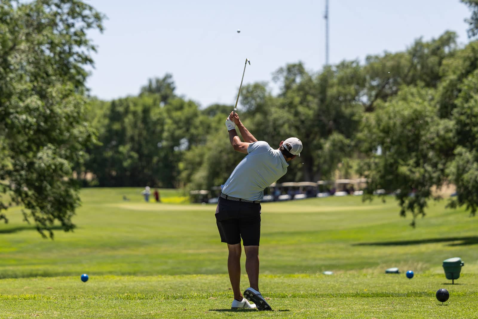 Golfer mid-swing on a sunny golf course, wearing light blue polo and black shorts, with a club raised above his head.