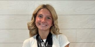 Smiling young woman with two medals on black ribbons around her neck, wearing a white shirt, against a light tiled wall.