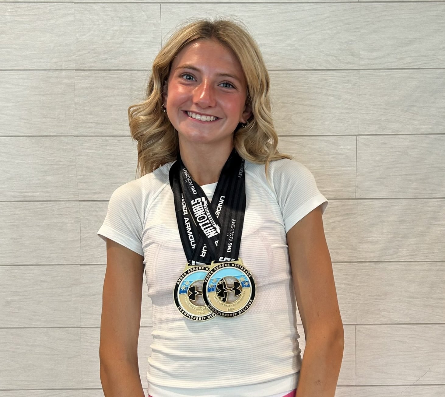 IMG_5232 | Press Pass Sports Smiling young woman with two medals on black ribbons around her neck, wearing a white shirt, against a light tiled wall.