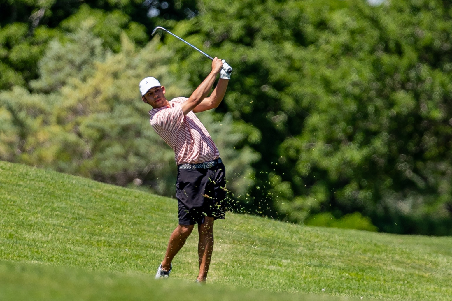 Golfer in a striped polo swinging a club on a grassy fairway, sending dirt and grass into the air as he follows through.