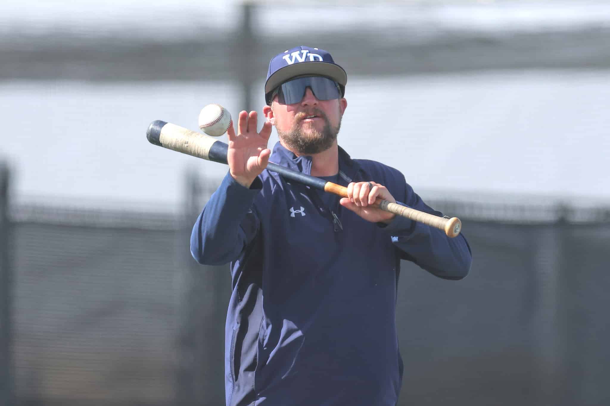 Baseball player in a navy jacket and cap with reflective sunglasses, holding a bat mid-swing as a baseball approaches.