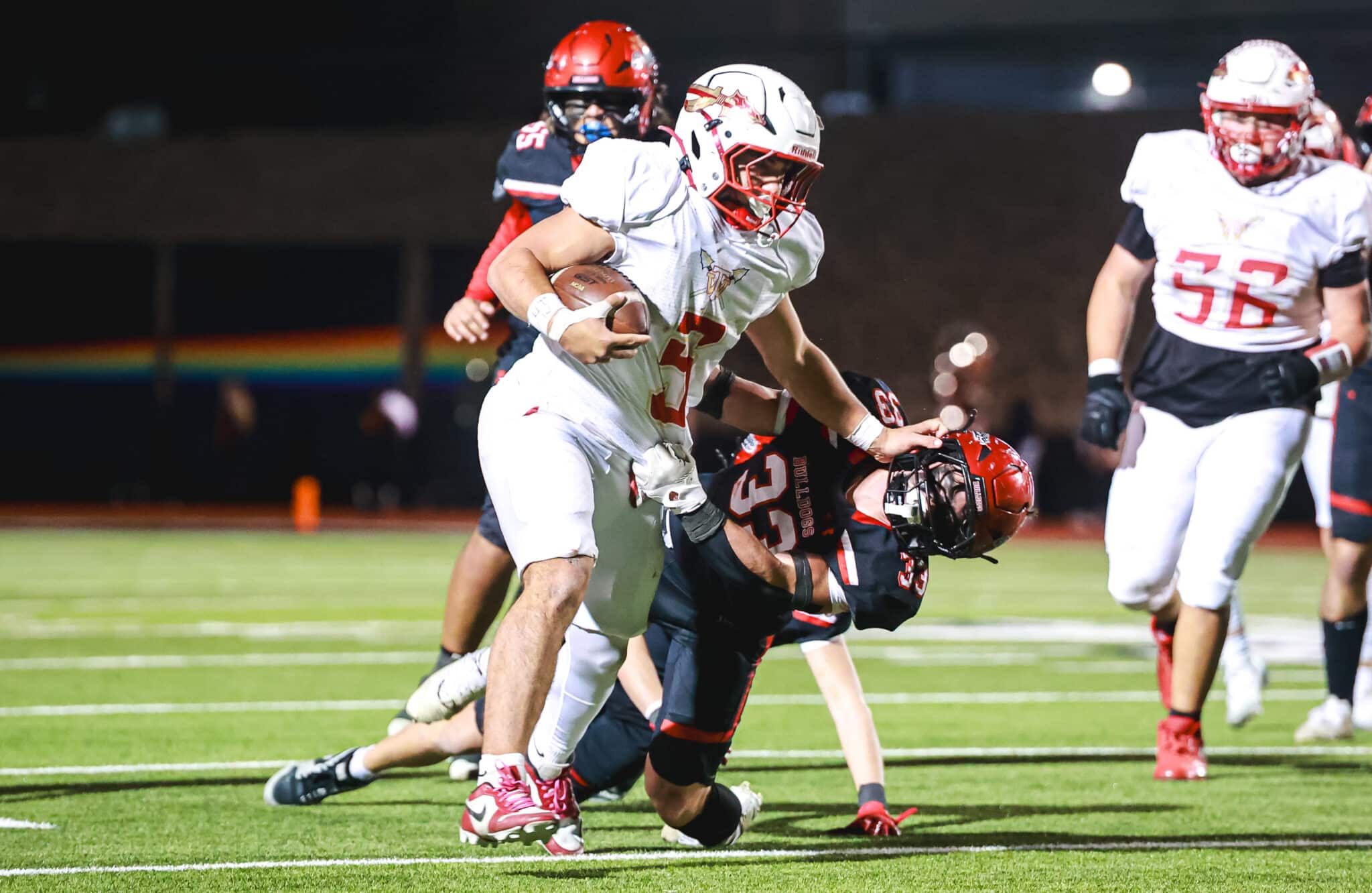 MY4A7364 1 | Press Pass Sports Football player in a white uniform runs with the ball as opponents in red/black jerseys tackle him on a green turf field at night, stadium lights in background.