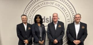 Four adults in business attire pose for a group photo in front of a large circular seal on the wall, all wearing dark blazers.