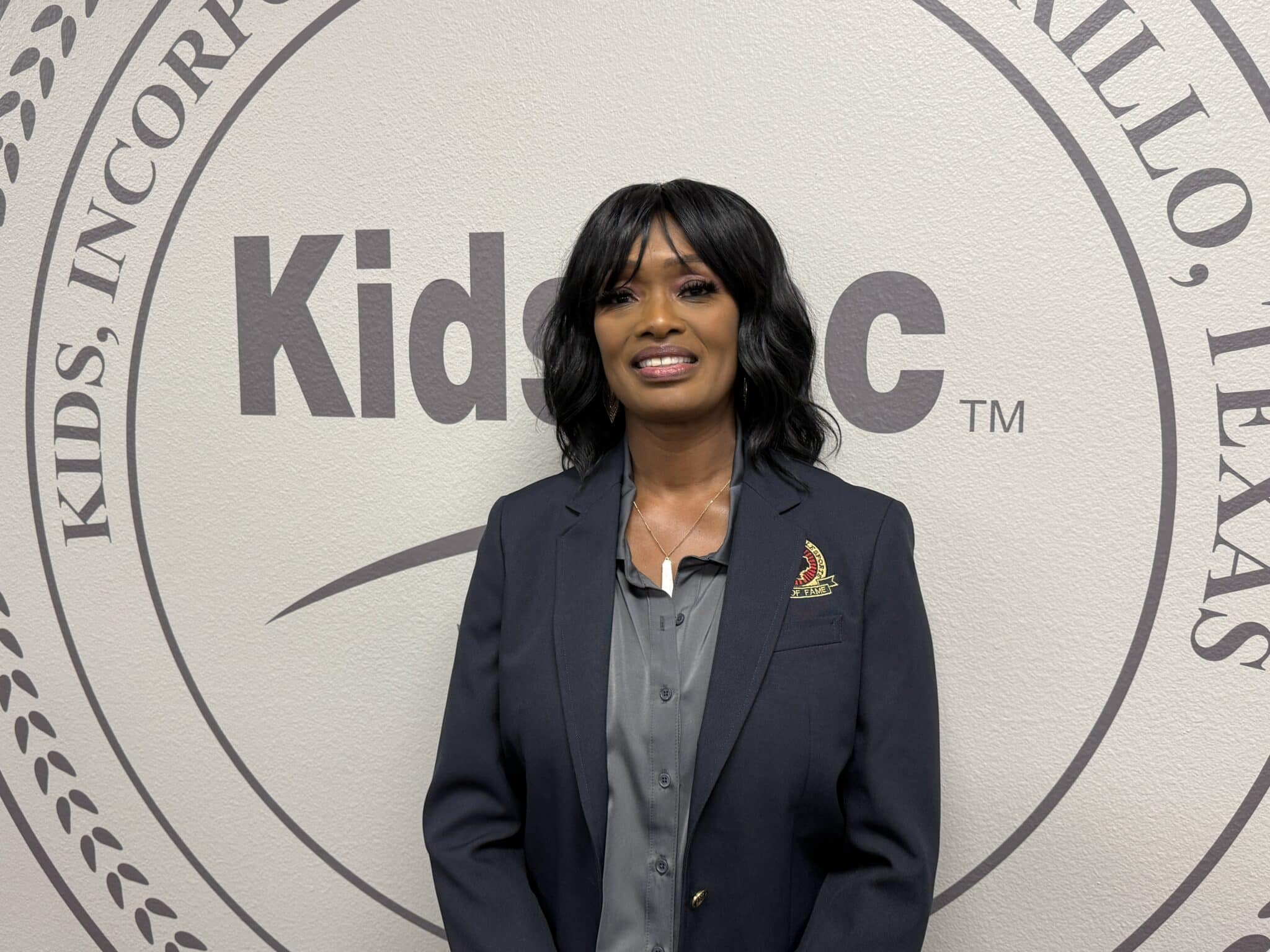 IMG_5306 | Press Pass Sports Professional woman in a navy blazer poses in front of a large wall seal with the word'Kids'.