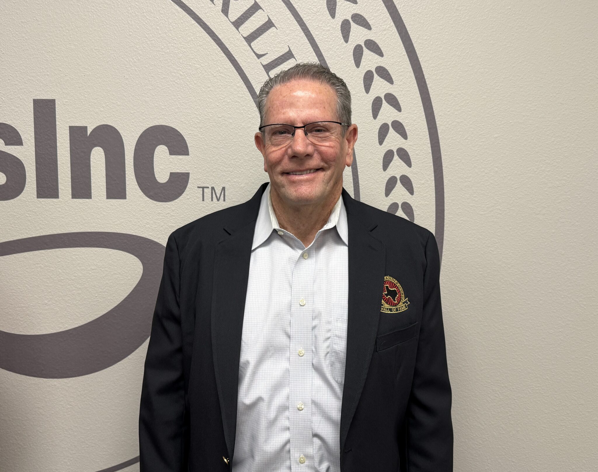 IMG_5303 | Press Pass Sports Smiling man in a dark suit jacket and light shirt posing in front of a beige wall with a large logo.
