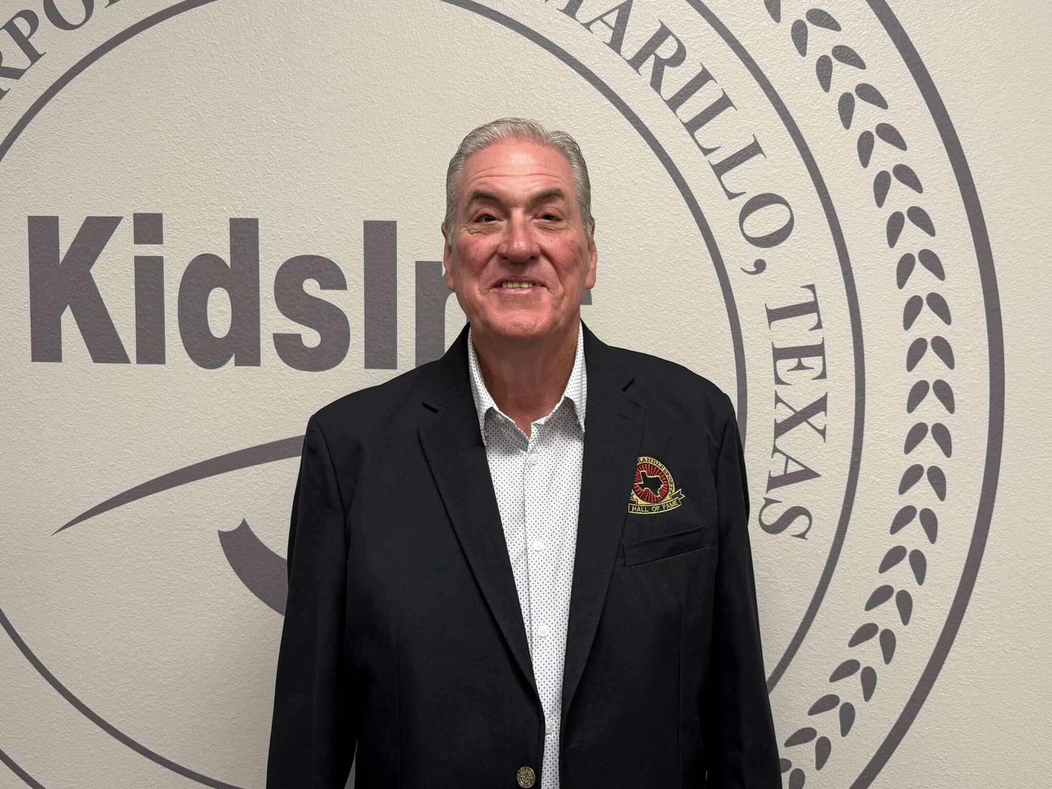 IMG_5299 | Press Pass Sports Smiling man in a dark suit stands in front of a large circular emblem on a beige wall behind him Appear to be in a formal setting or event backdrop.